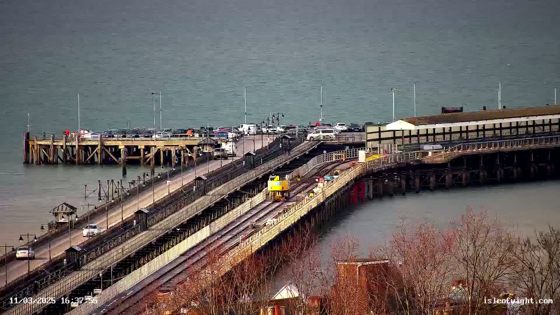 A long pier extends over calm, gray water, with a yellow vehicle on its railway tracks, cars parked along its roadway, and bare trees visible in the foreground.