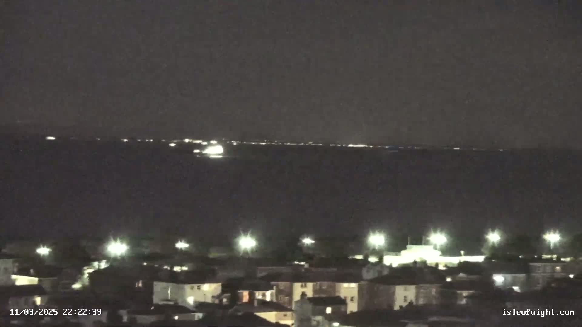 A nighttime view shows a dark body of water with faint lights on the horizon and a town with illuminated buildings and streetlights in the foreground under a dark sky.