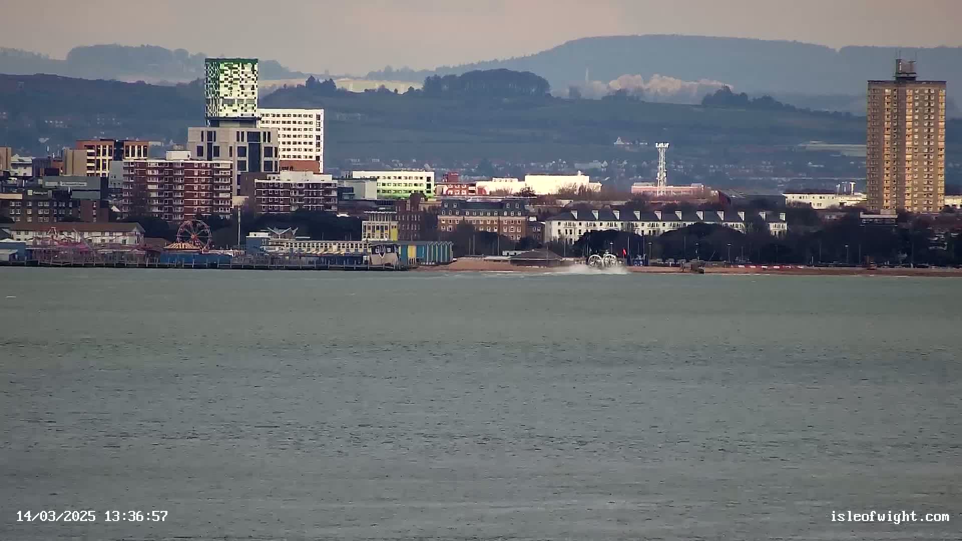 A seaside town with various buildings, including high-rises and a Ferris wheel, is seen across a calm body of water under an overcast sky.