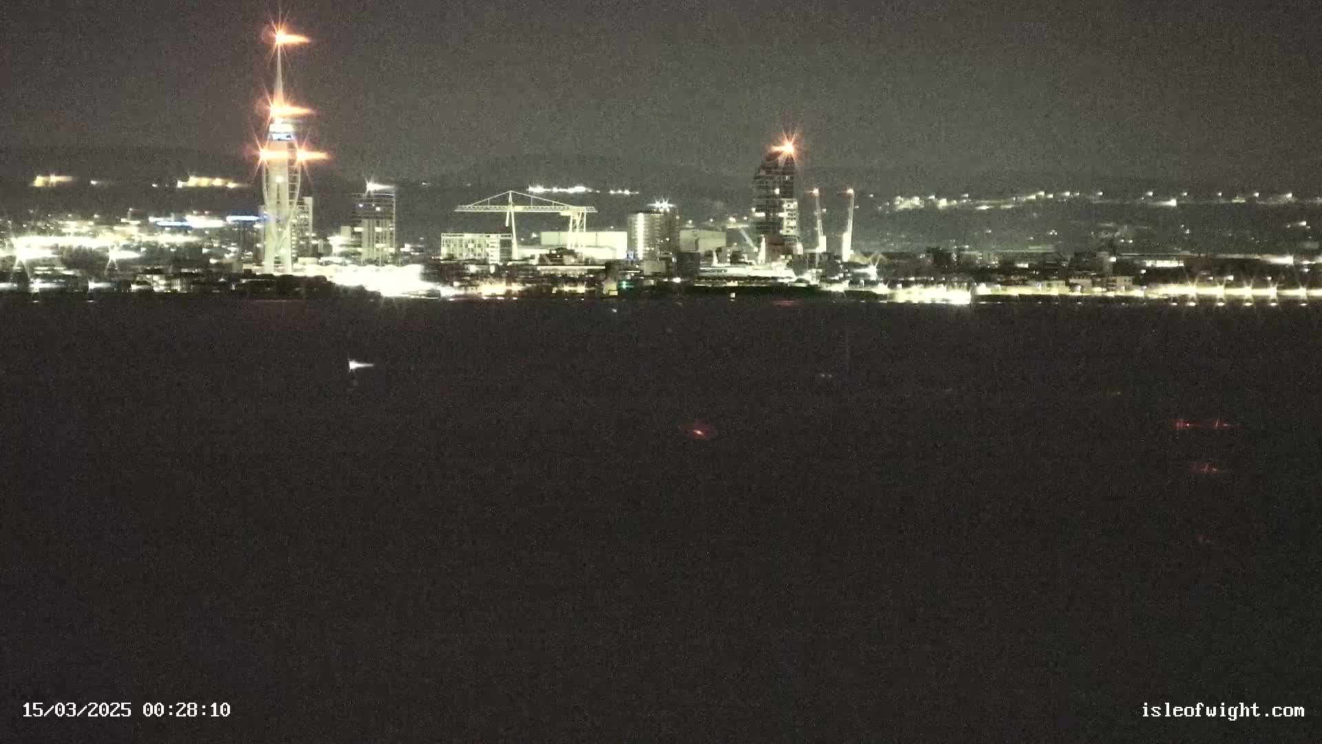 A nighttime view of a city skyline across a dark body of water, featuring illuminated buildings and towers.