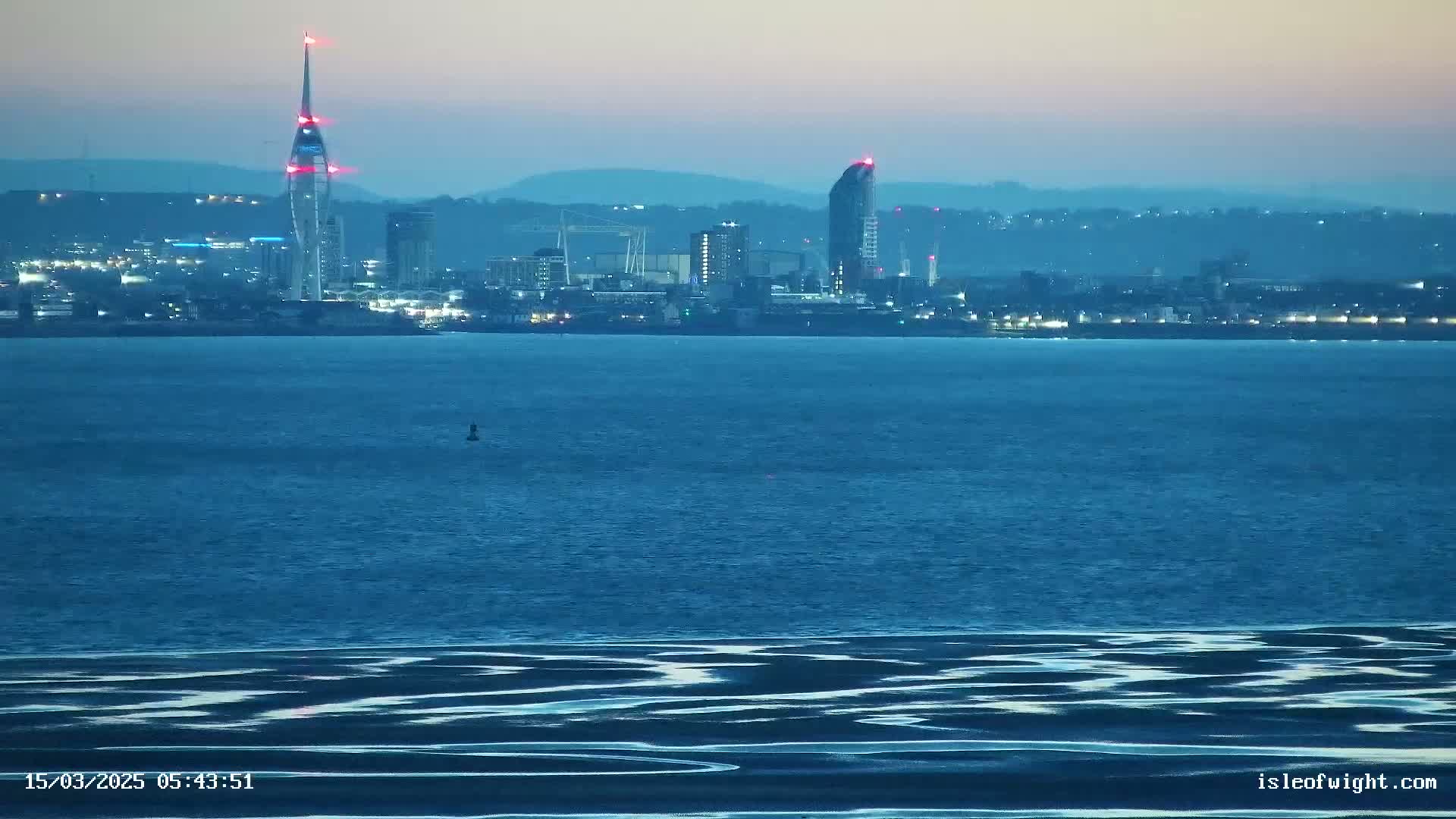 A city skyline with a tall, slender tower and a high-rise building is visible across a body of dark-blue water under a twilight sky.