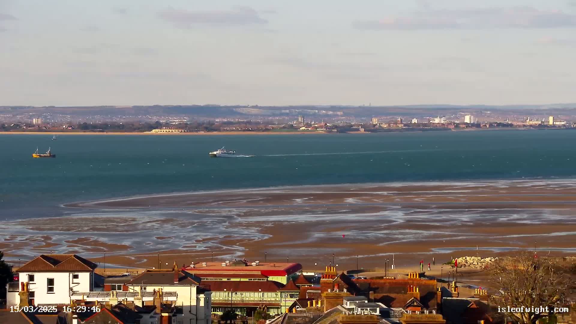 A coastal town overlooks a wide expanse of teal water with exposed sandbars at low tide, a distant cityscape visible in the background under a clear sky.