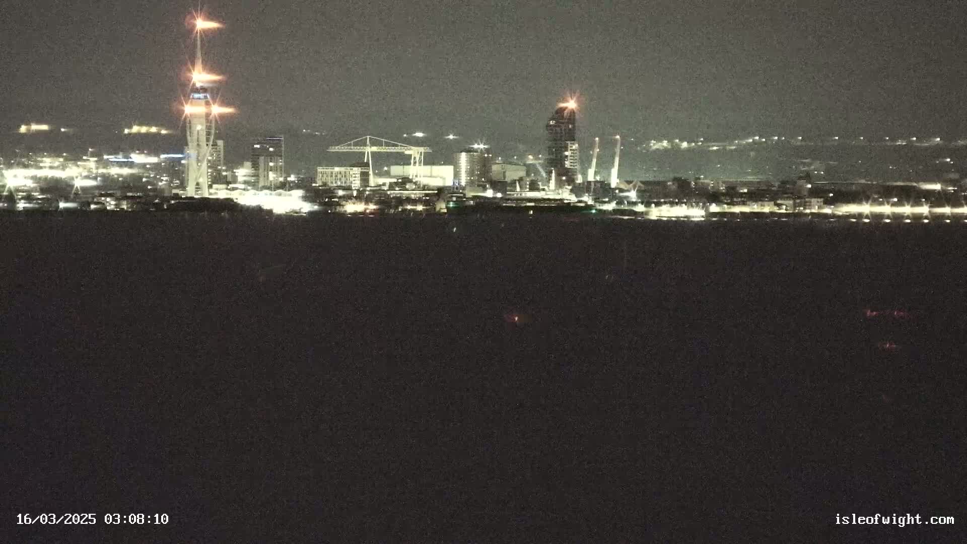 A nighttime view of a city skyline across a dark body of water, featuring illuminated buildings and a tall tower.
