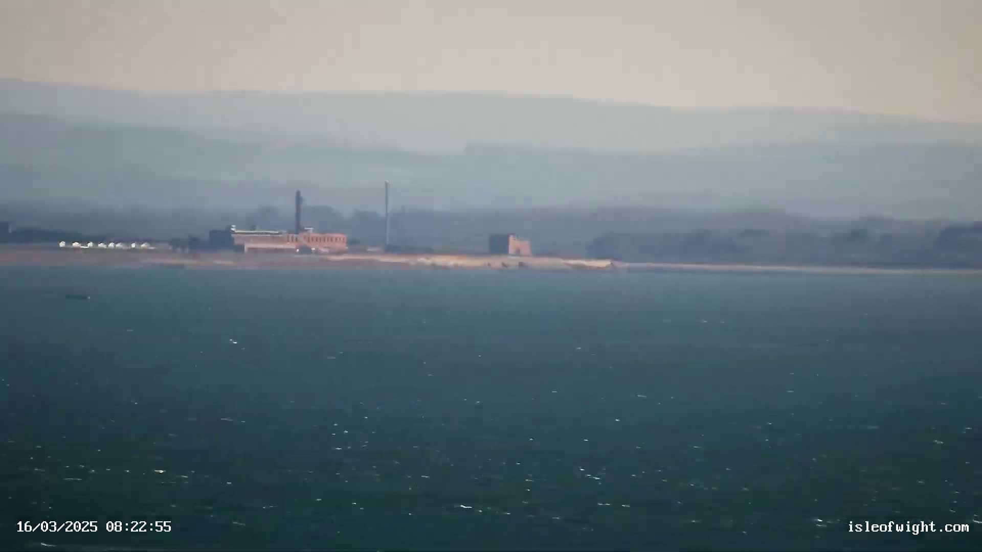 A hazy view shows a distant shoreline with buildings and a tall structure, all situated across a body of somewhat choppy water.