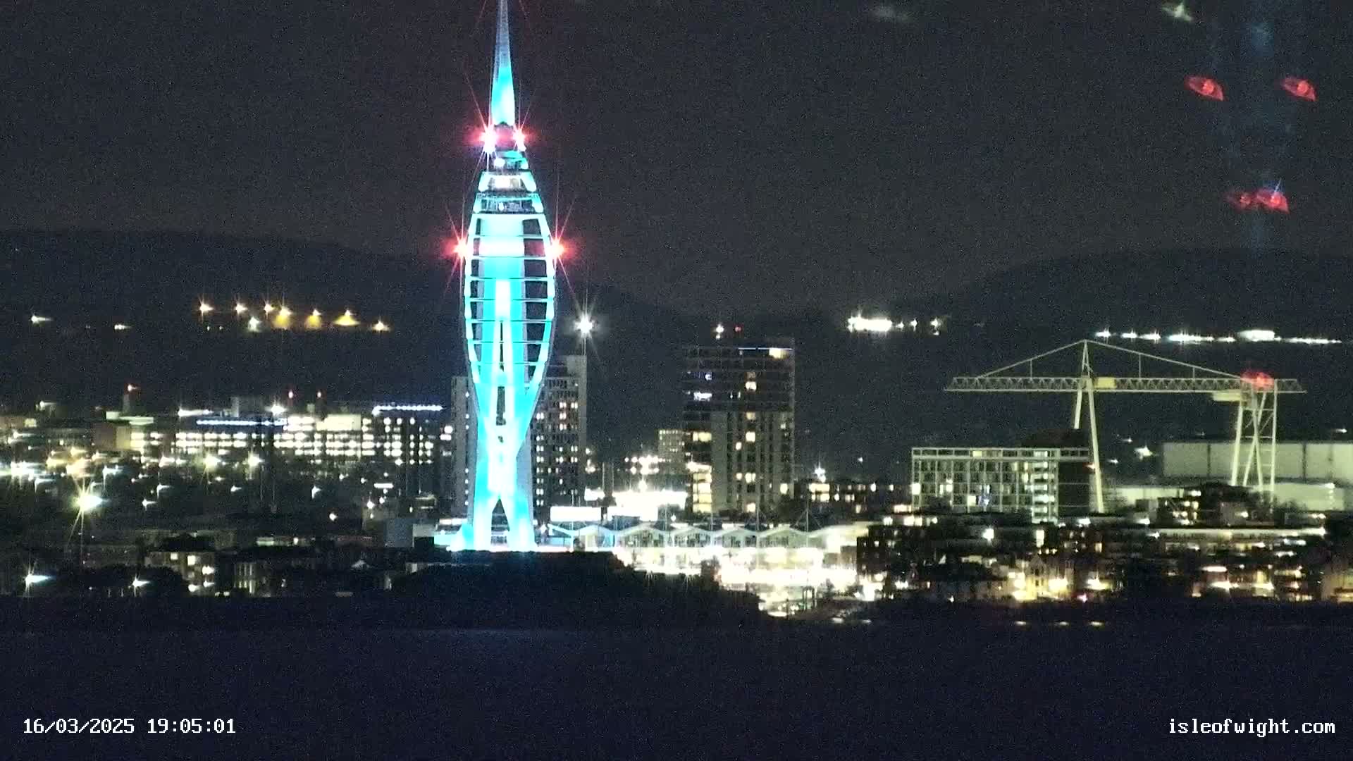 A brightly lit, tall, slender tower stands out against a dark night skyline of a city with buildings and a crane illuminated by lights, all viewed from across a body of water.