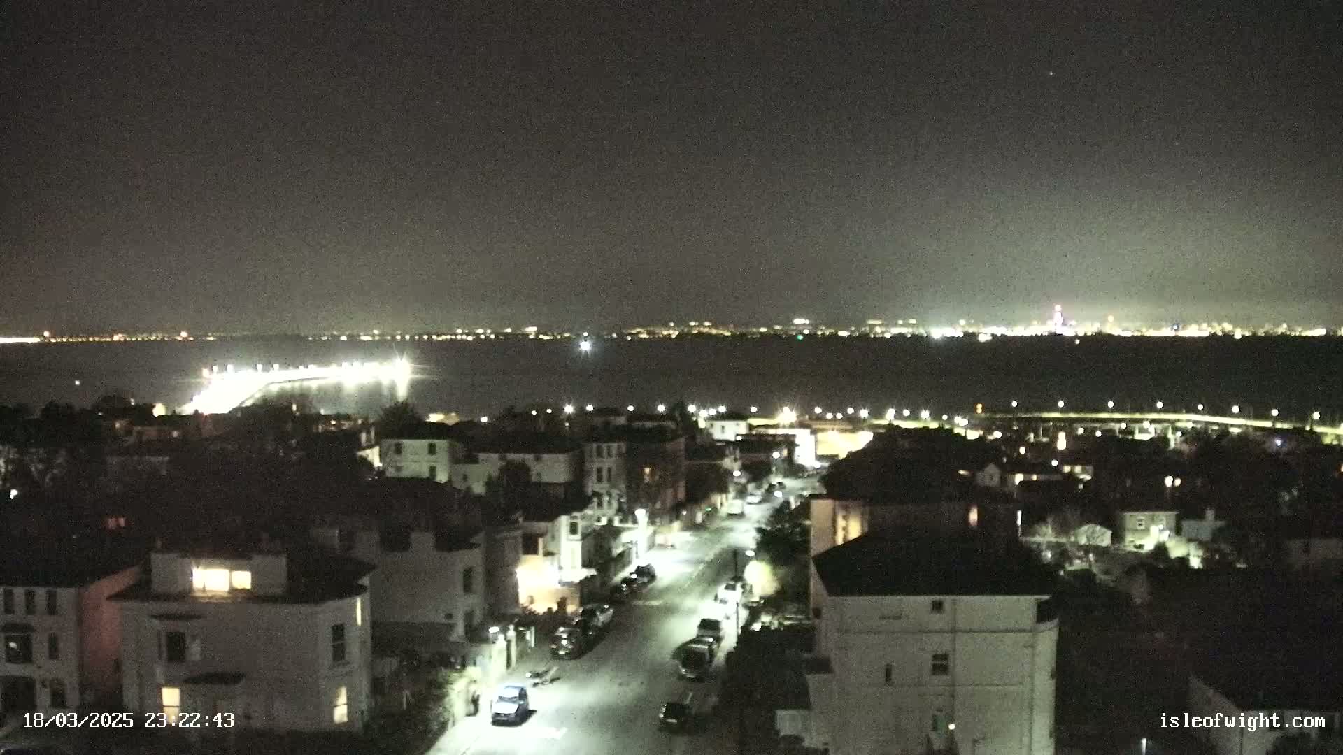 A nighttime view of a residential street illuminated by streetlights, overlooking a body of water with a distant city skyline visible under a clear, dark sky.