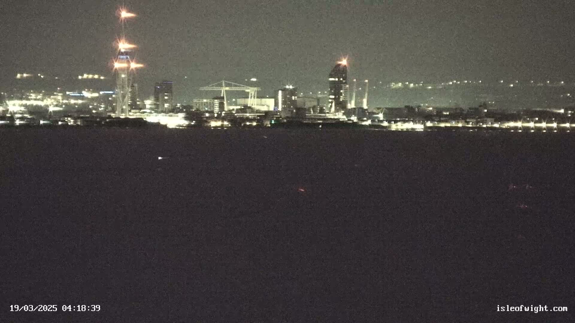 A nighttime view of a city skyline across a dark body of water, featuring several illuminated buildings and a tall tower with bright lights at its peak.