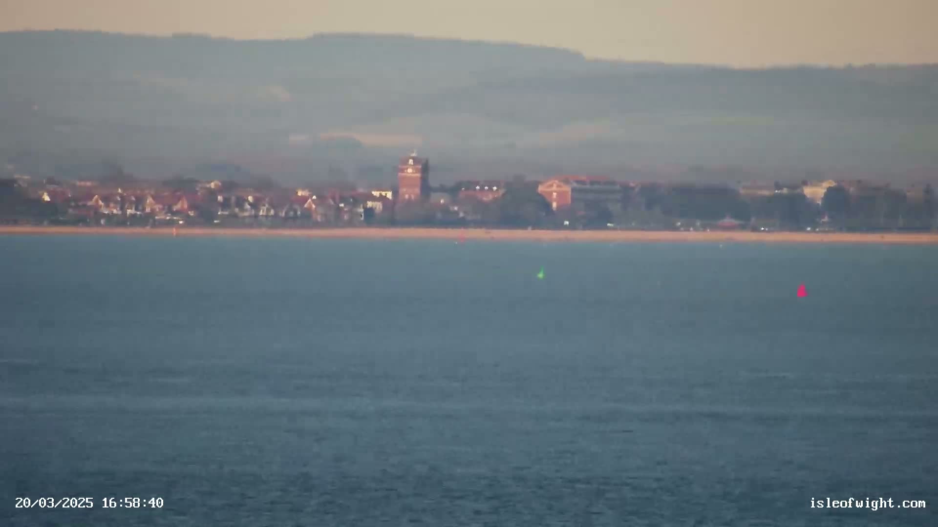 A hazy coastal town with a prominent tower is visible across a calm body of water, with two small boats in the distance.