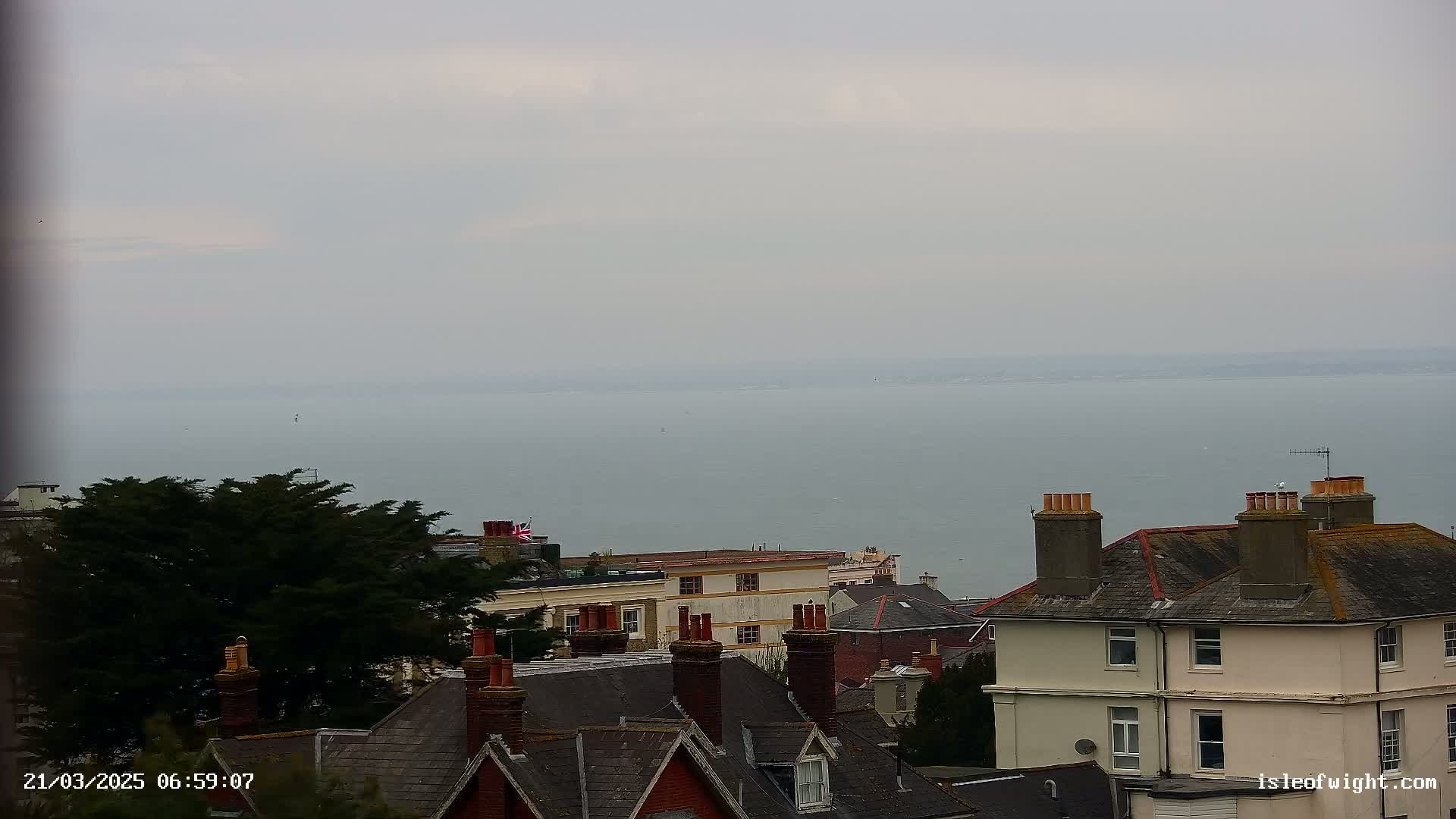 A calm, overcast day reveals a view of a body of water beyond a collection of houses with dark gray roofs and brick chimneys.