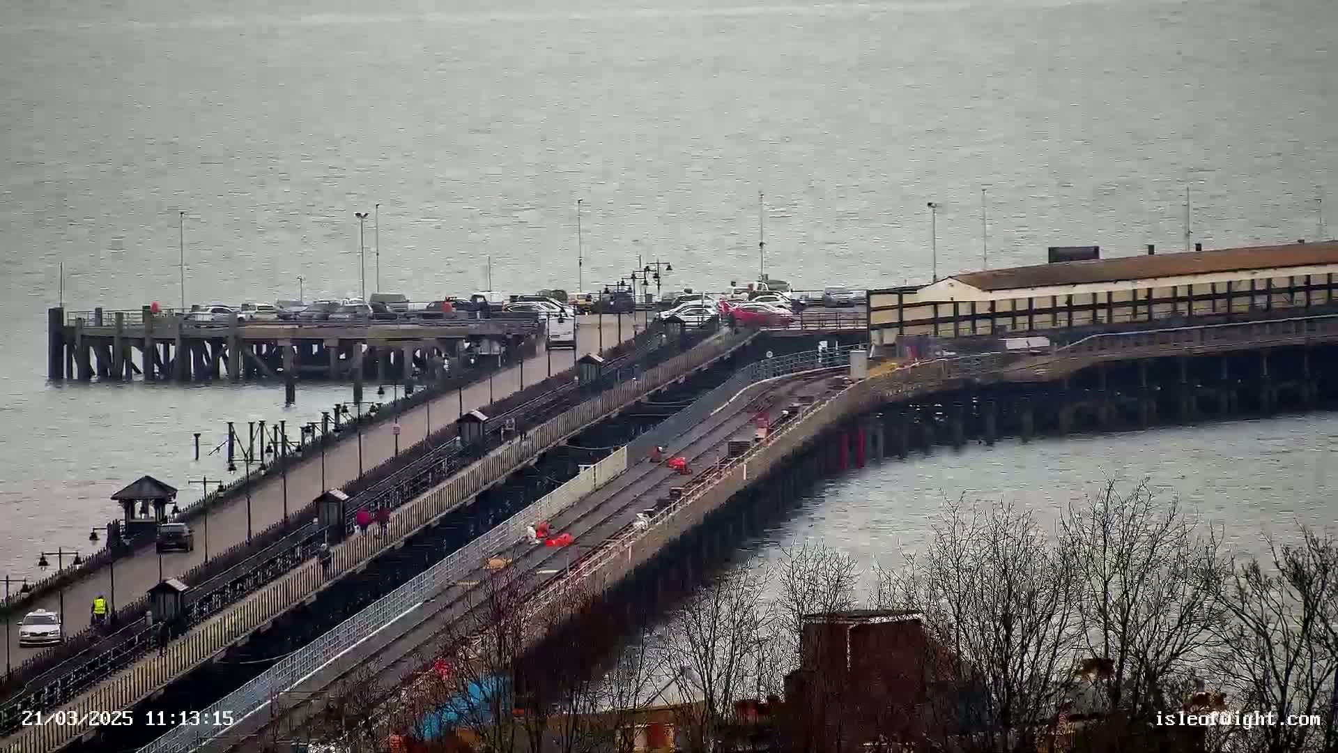 A long pier extends over a calm, gray body of water, with cars parked on a section of the pier and people walking along its walkway on an overcast day.