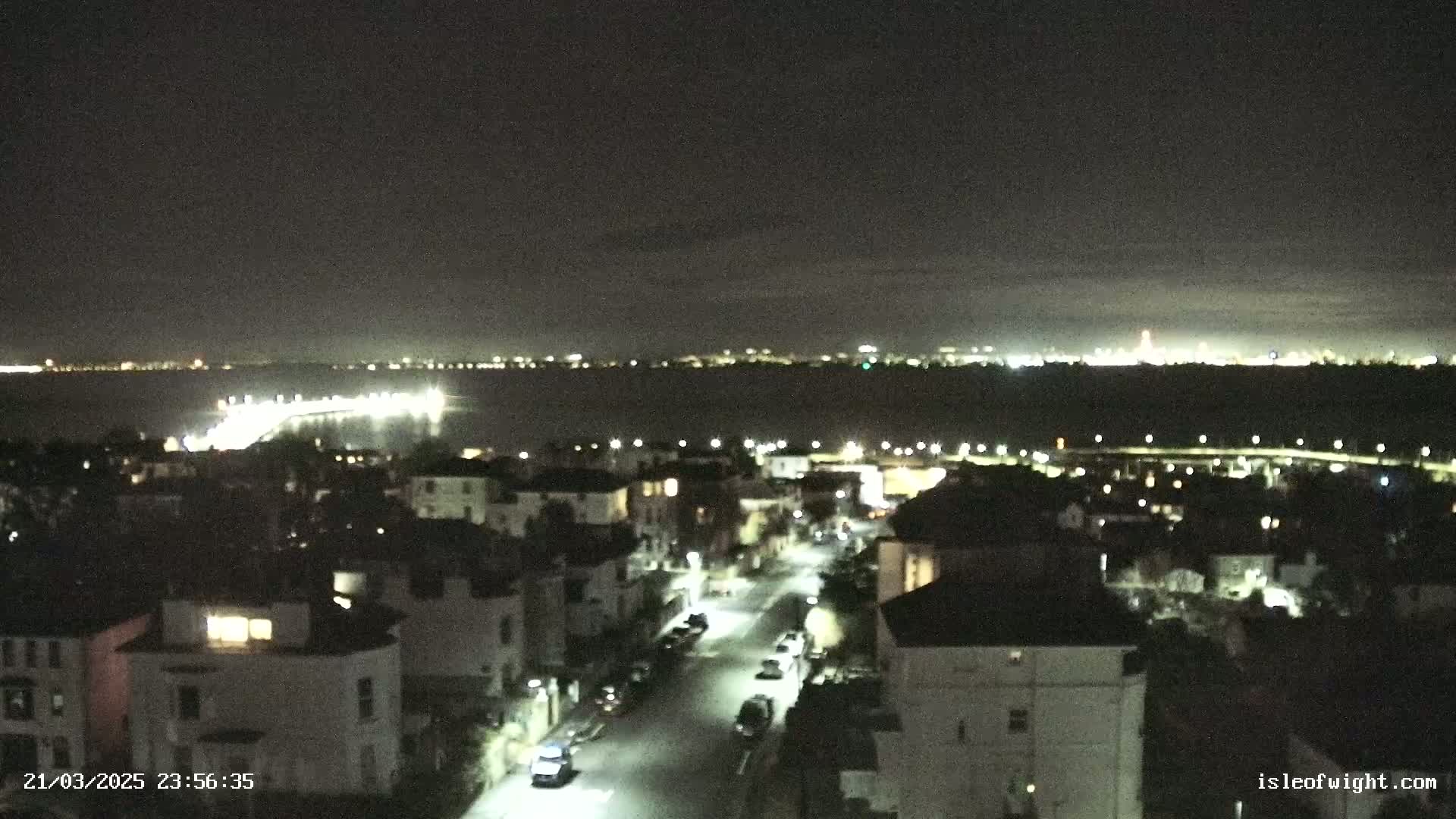 A nighttime view shows a city street with parked cars, illuminated houses, and a body of water with lights across the horizon under a dark, clear sky.