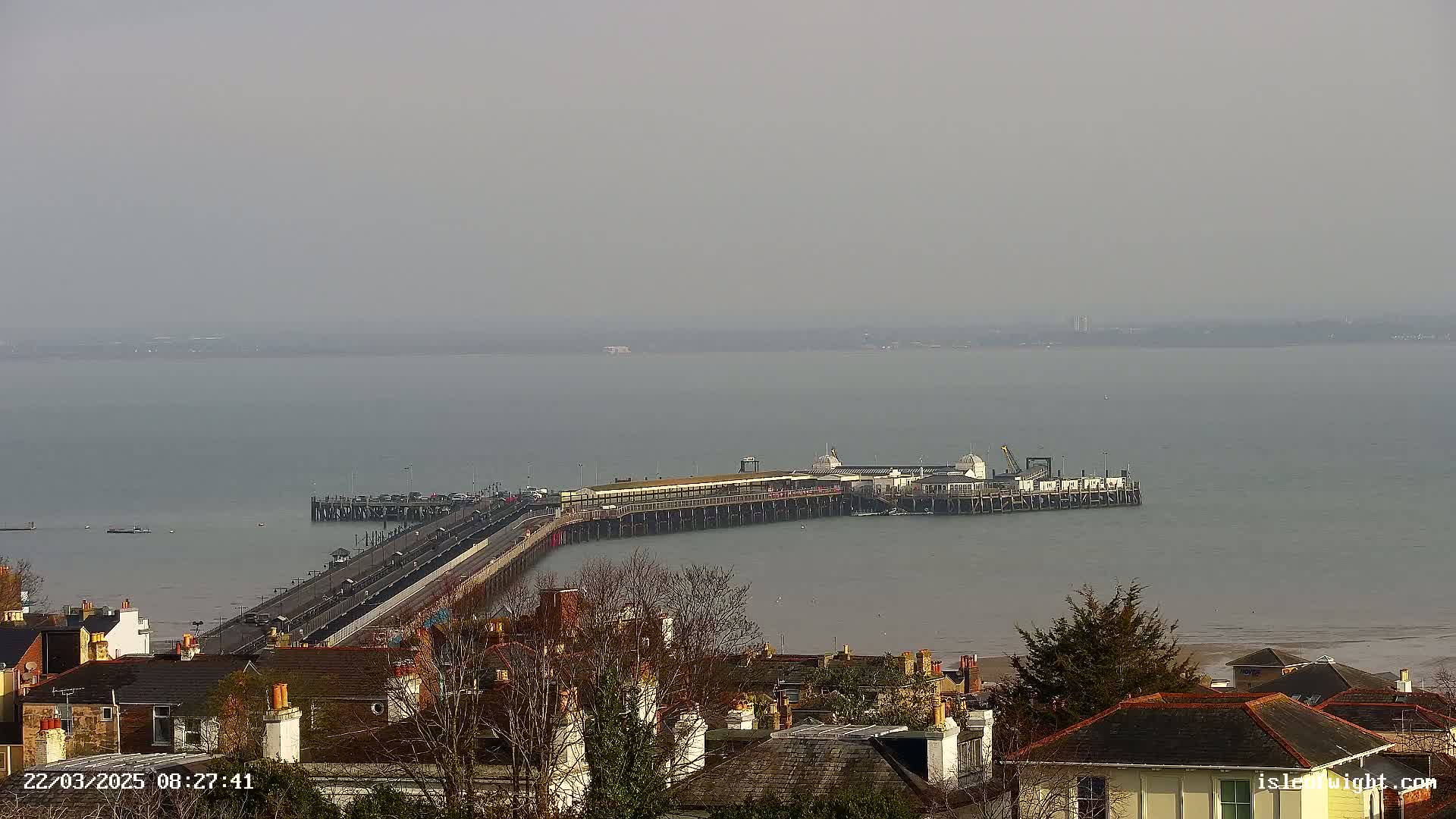 A long pier extends into a calm, hazy body of water, viewed from an elevated position overlooking a town of rooftops.