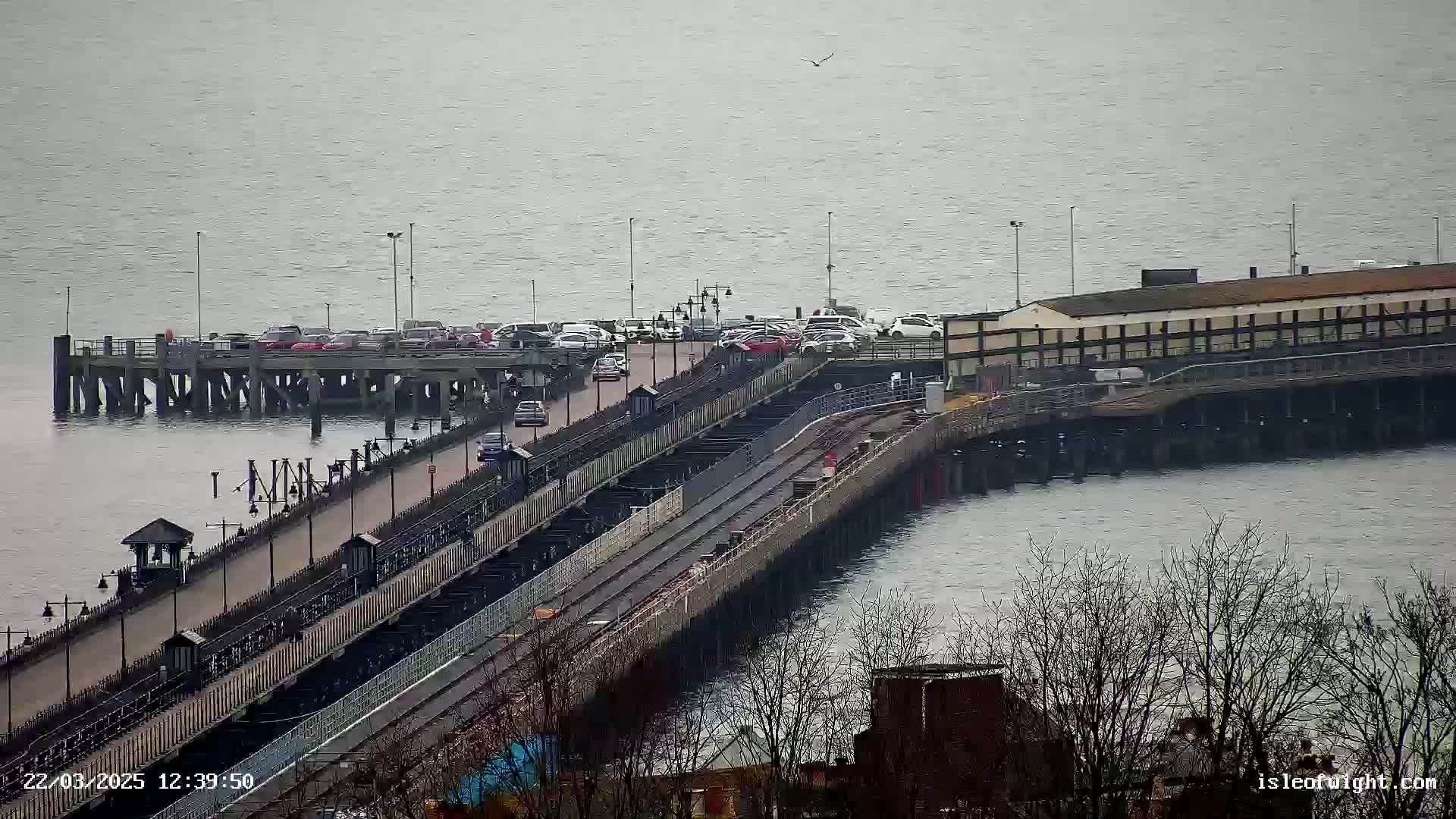 A long pier extends into a calm, gray body of water, with cars parked on a section of the pier and a few vehicles traveling along its roadway.