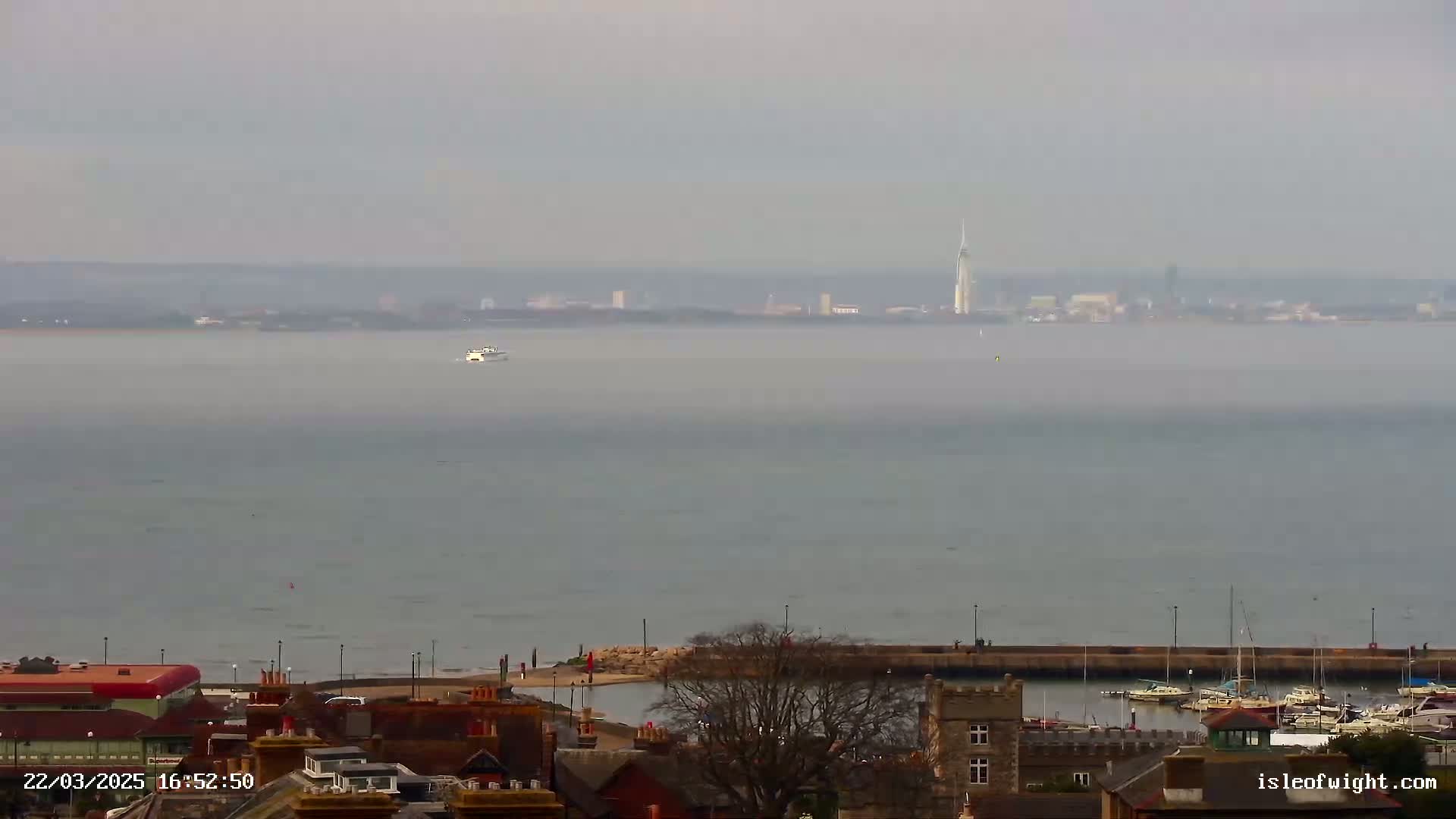 A hazy view of a body of water shows a distant city skyline with a tall tower, a small boat on the water, and a small harbor with boats in the foreground.