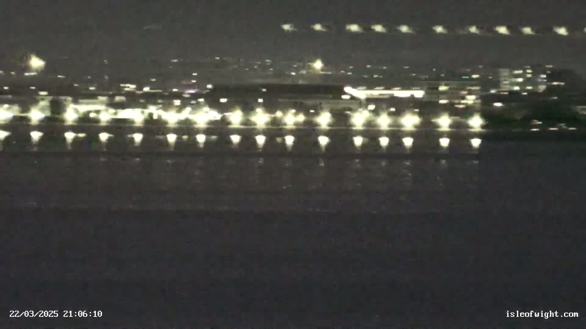 A nighttime long shot shows a body of dark water reflecting the lights of a distant city and a line of lights closer to the viewer, possibly on a bridge or pier.