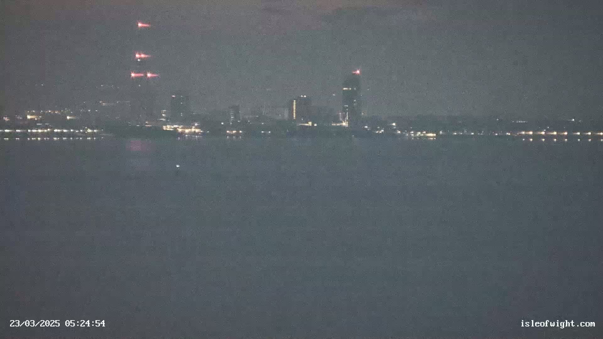 A nighttime view across a dark body of water shows a distant city skyline with several lit buildings and three reddish lights possibly indicating construction or other activity.