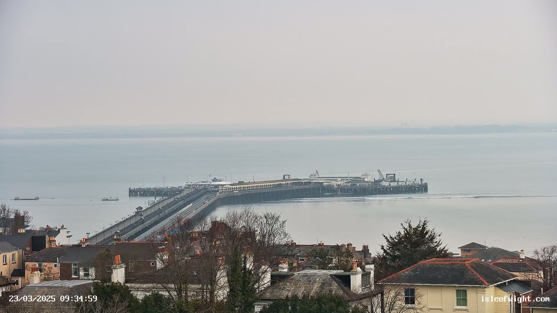 A long pier extends into a calm, gray body of water, viewed from a hillside overlooking a town of houses with gray roofs.