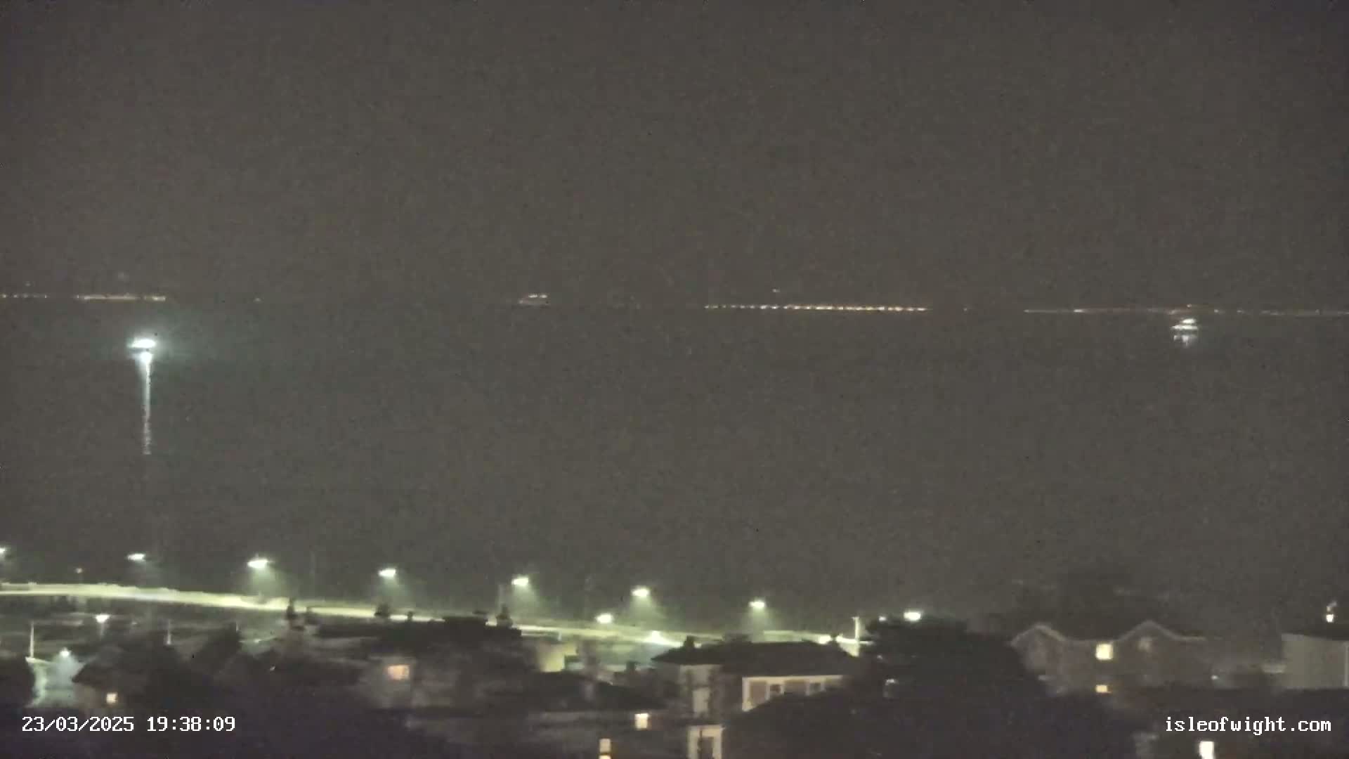A nighttime view of a dark body of water with a distant line of lights, a bright light on a pole in the water, and a shoreline with dimly lit buildings and streetlights.