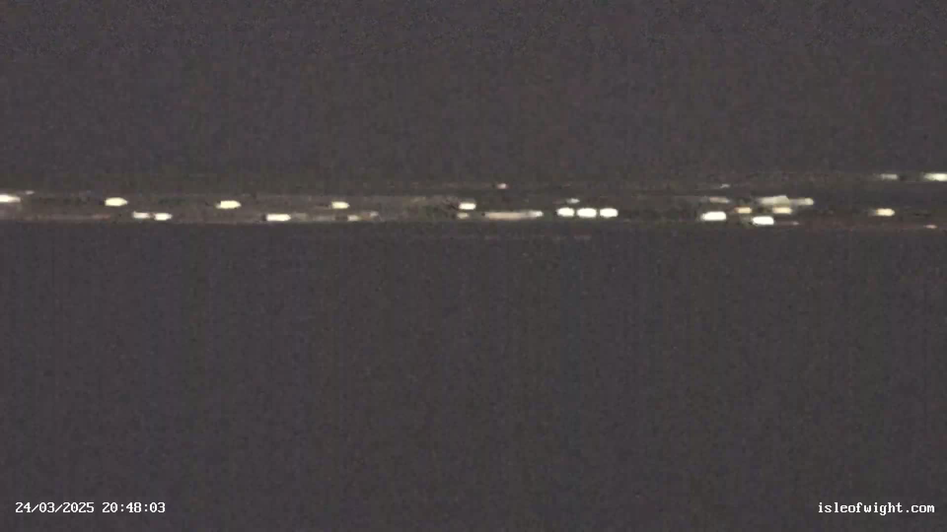 A dark, nighttime scene shows a distant line of lights across a dark body of water under a clear, dark sky.
