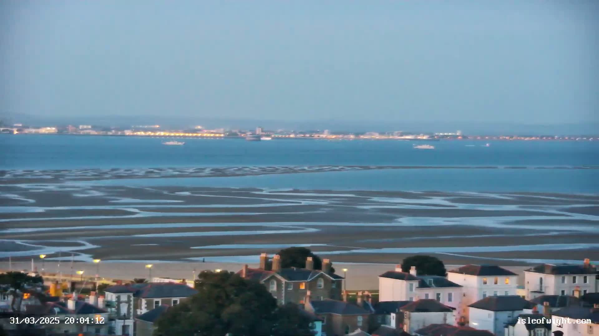 A coastal town sits atop a hill overlooking a wide expanse of tidal flats and a calm body of water with a distant city skyline under a twilight sky.