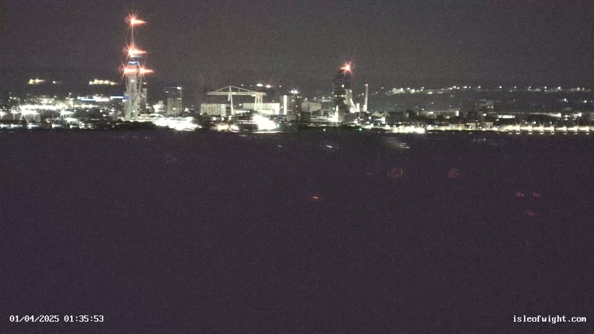 A nighttime view of a city skyline across a dark body of water, featuring illuminated buildings and a tall tower.