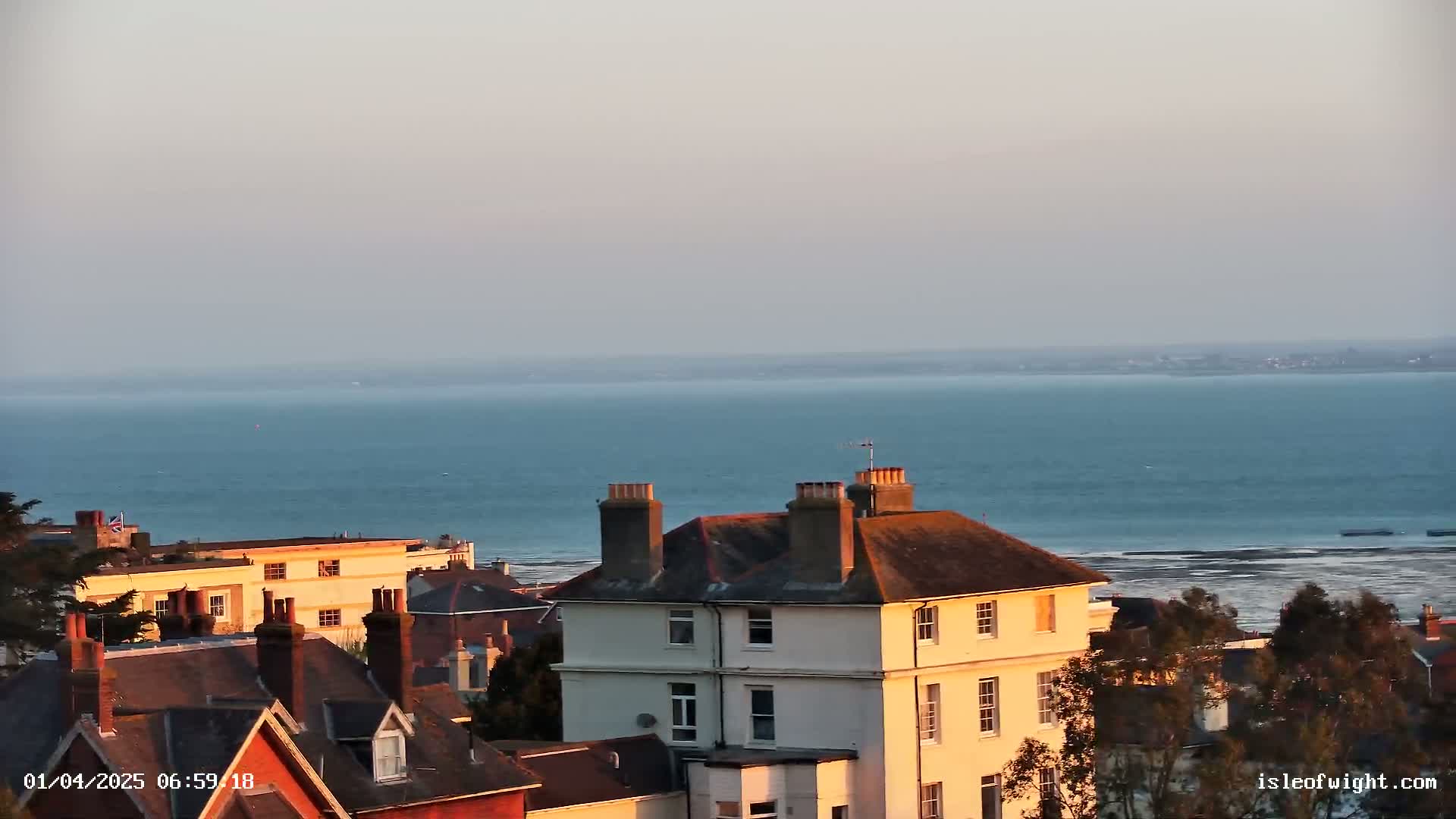 A calm, sunlit ocean stretches beyond a collection of houses with red-tiled roofs and chimneys.