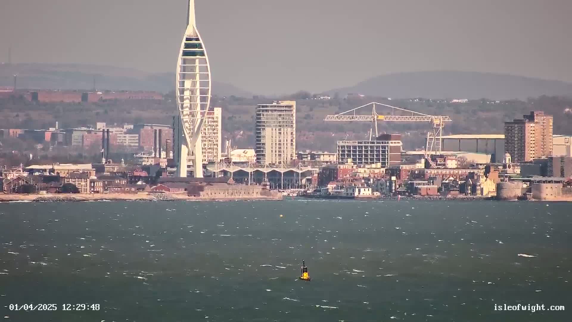A city skyline with a tall, slender tower is visible across a choppy body of water under a hazy sky.