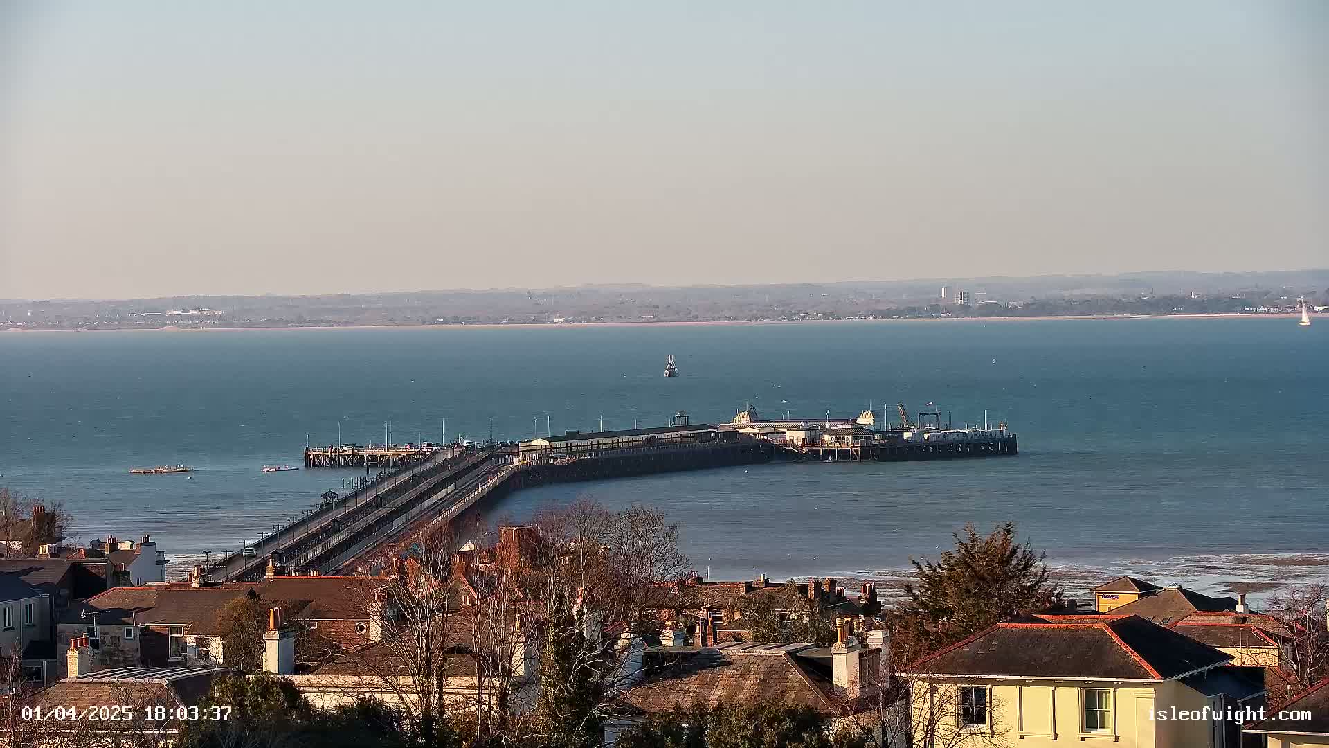 A long pier extends into a calm, blue body of water under a clear, light-blue sky, with a town visible in the foreground.