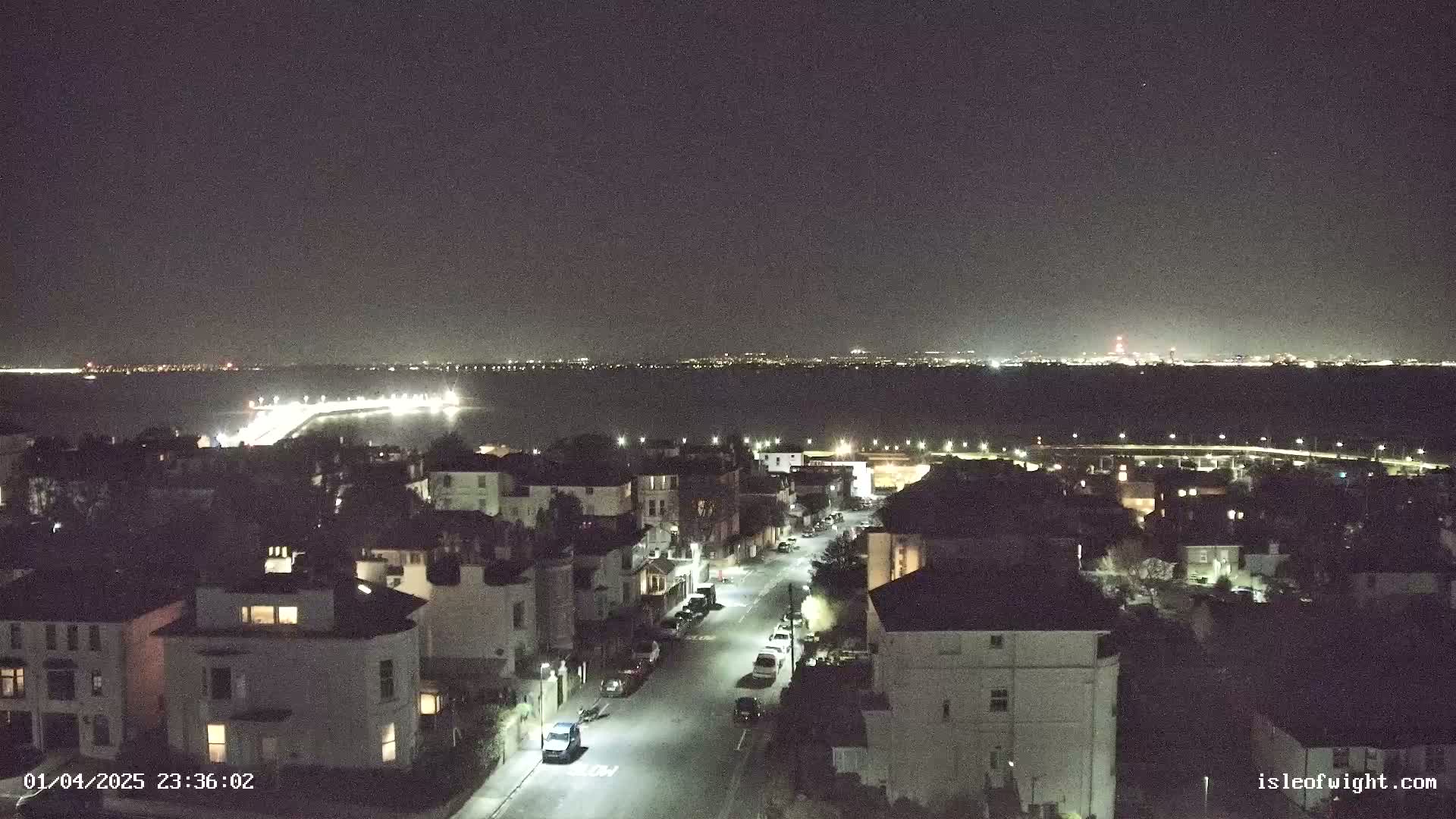 A nighttime, high-angle view shows a residential street illuminated by streetlights, overlooking a calm body of water with distant city lights under a clear, dark sky.