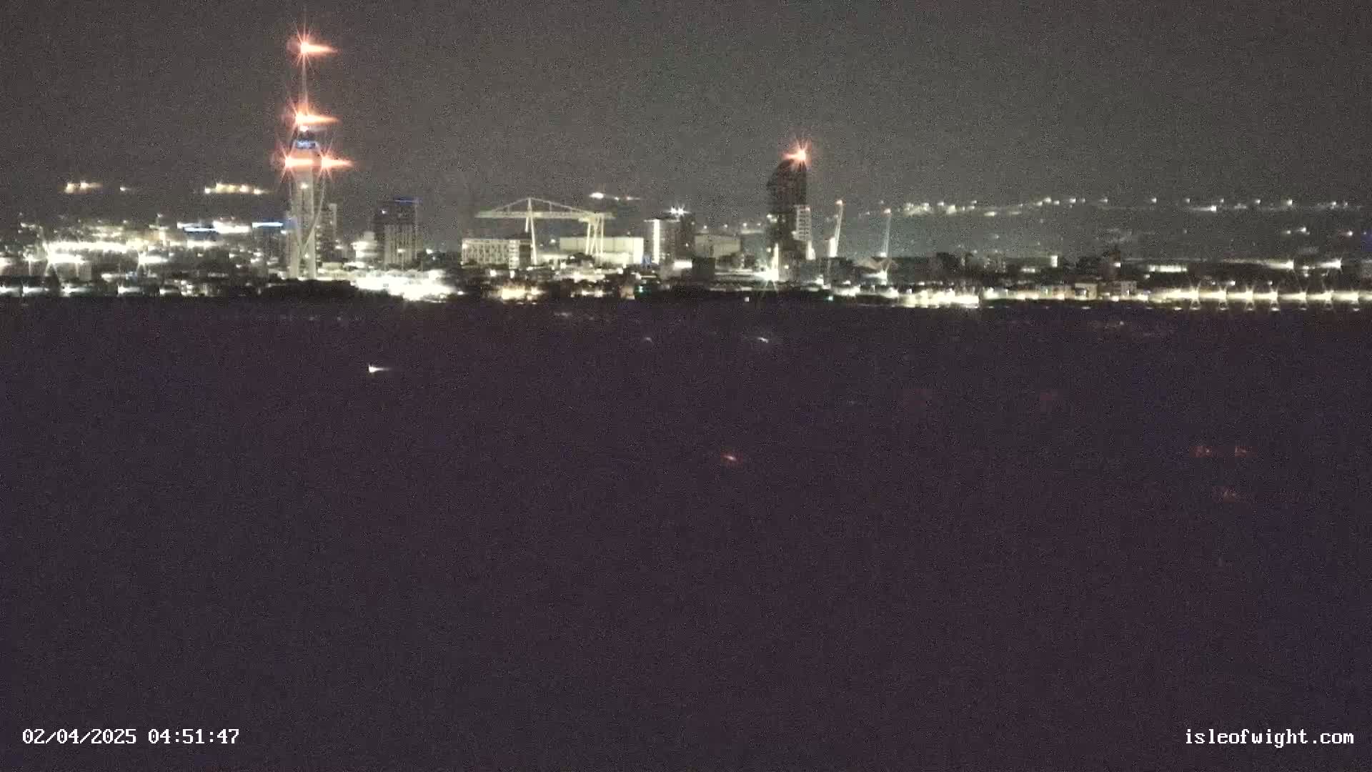 A nighttime view of a city skyline across a dark body of water, showing illuminated buildings, cranes, and tall structures.