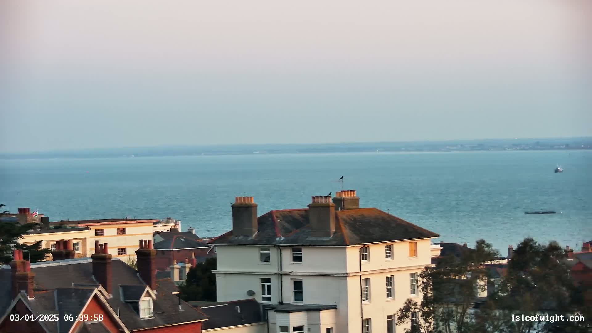 A seaside town's rooftops and a calm body of water stretch to the horizon under a pale sky.