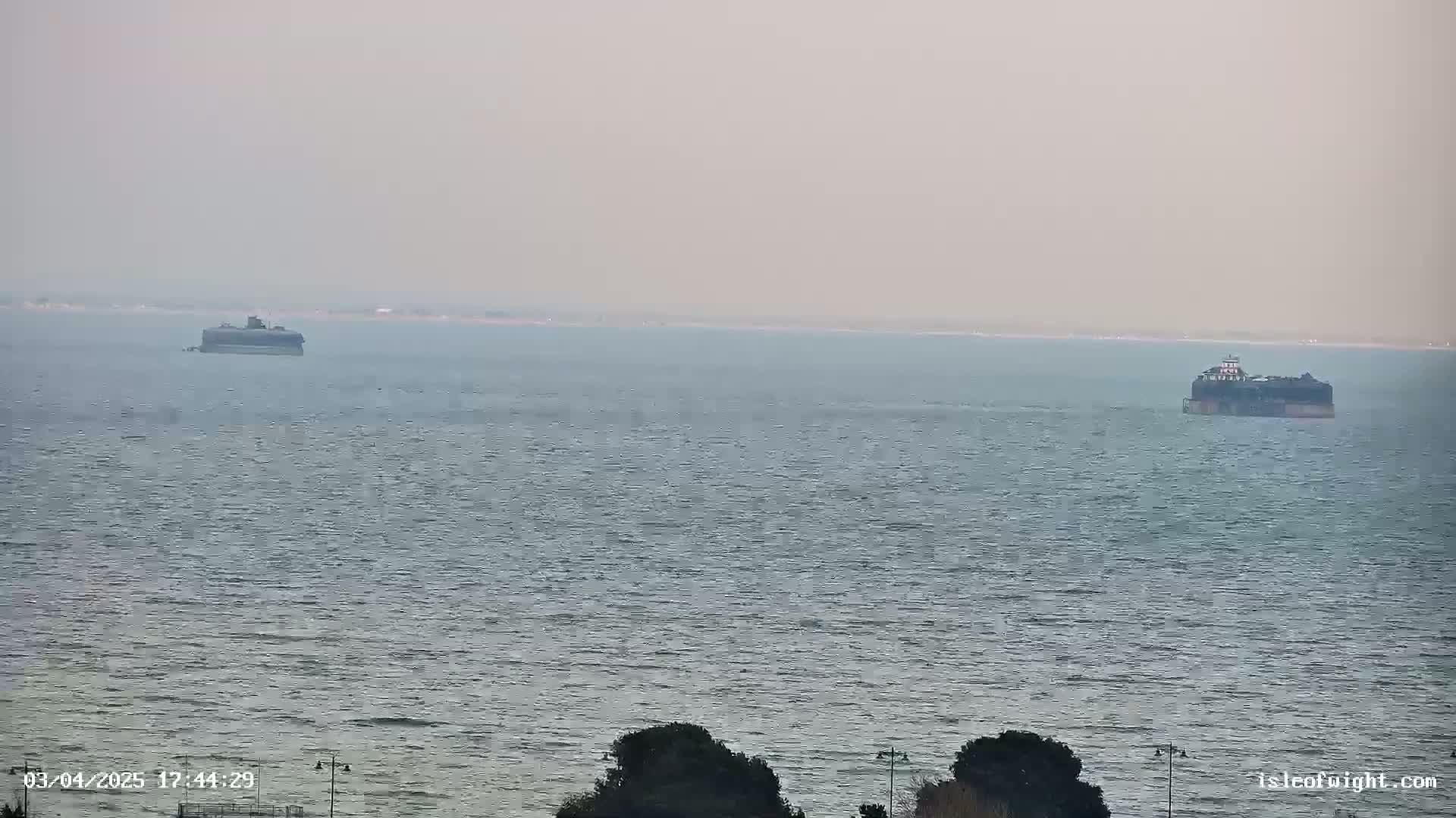 Two dark-colored structures sit on a calm, hazy ocean, with dark green vegetation visible in the bottom right corner.