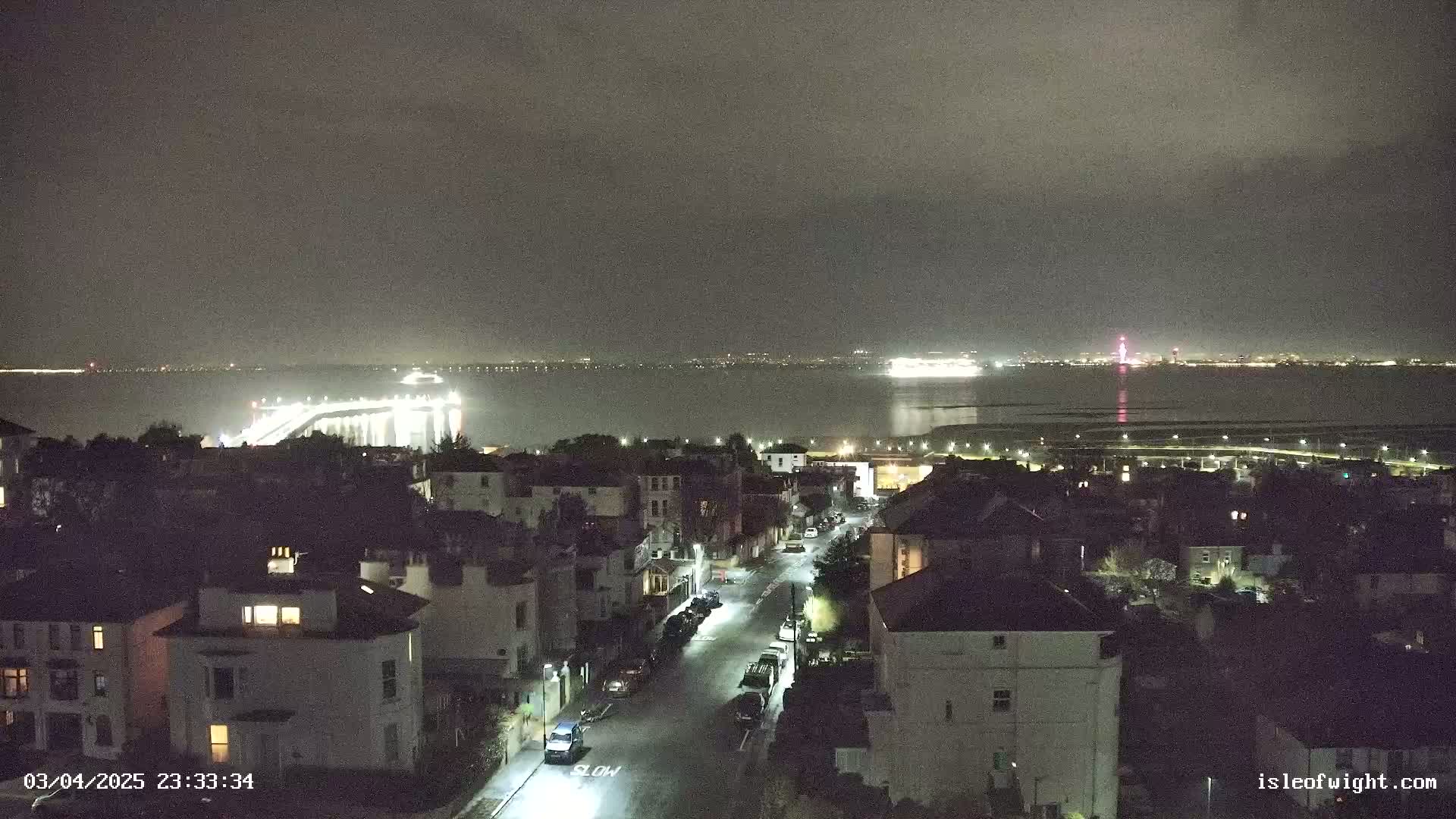 A nighttime view of a town overlooking a calm body of water, with a lit bridge and distant city lights visible under a cloudy sky.