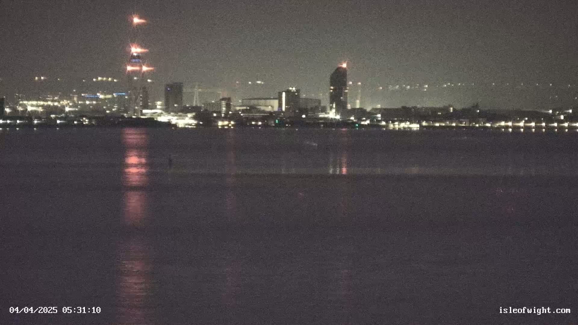 A nighttime view across a calm body of water shows a city skyline with several lit buildings and construction cranes, reflecting faintly on the water's surface.