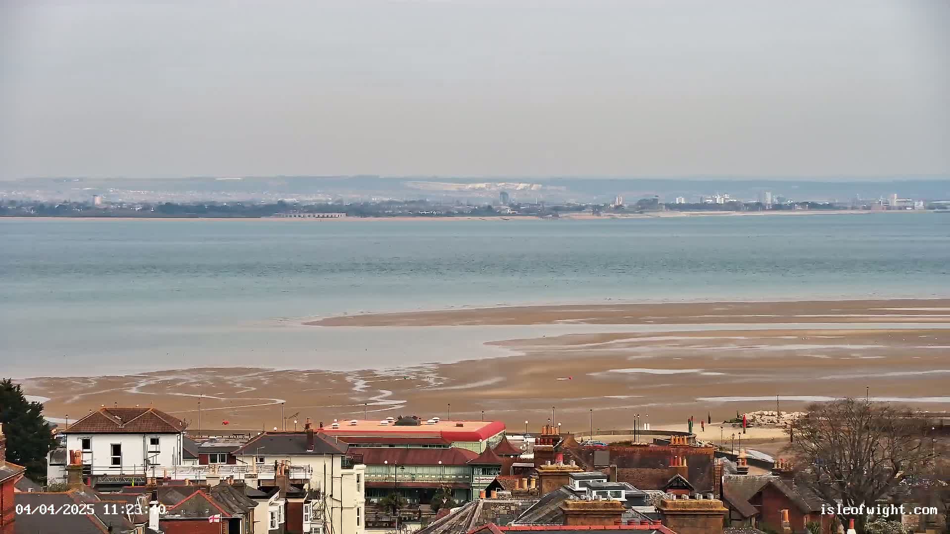 A coastal town overlooks a wide expanse of calm, light-blue water and exposed, muddy sandflats, with a distant city skyline visible on the horizon under an overcast sky.