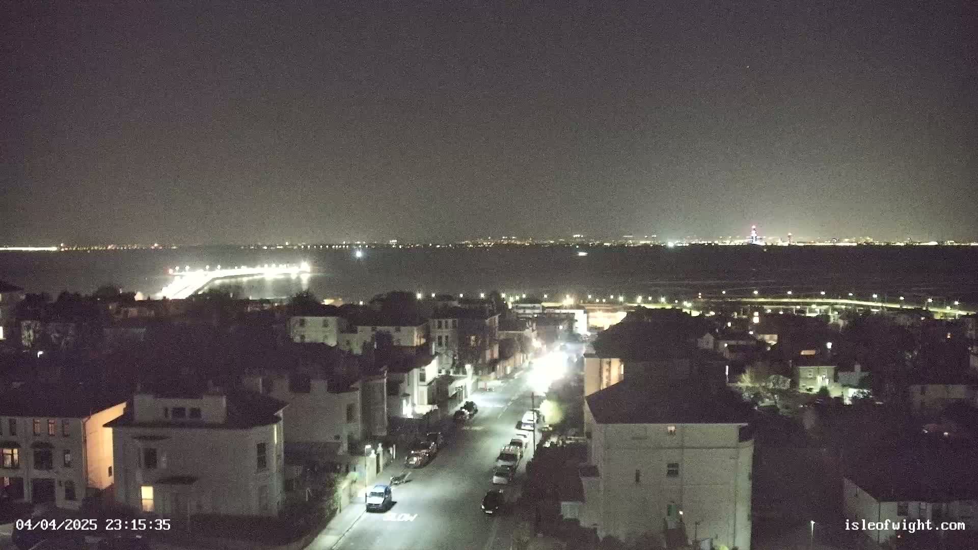 A nighttime, aerial view shows a residential street with parked cars, leading down toward a body of water with lights on the opposite shore under a clear, dark sky.