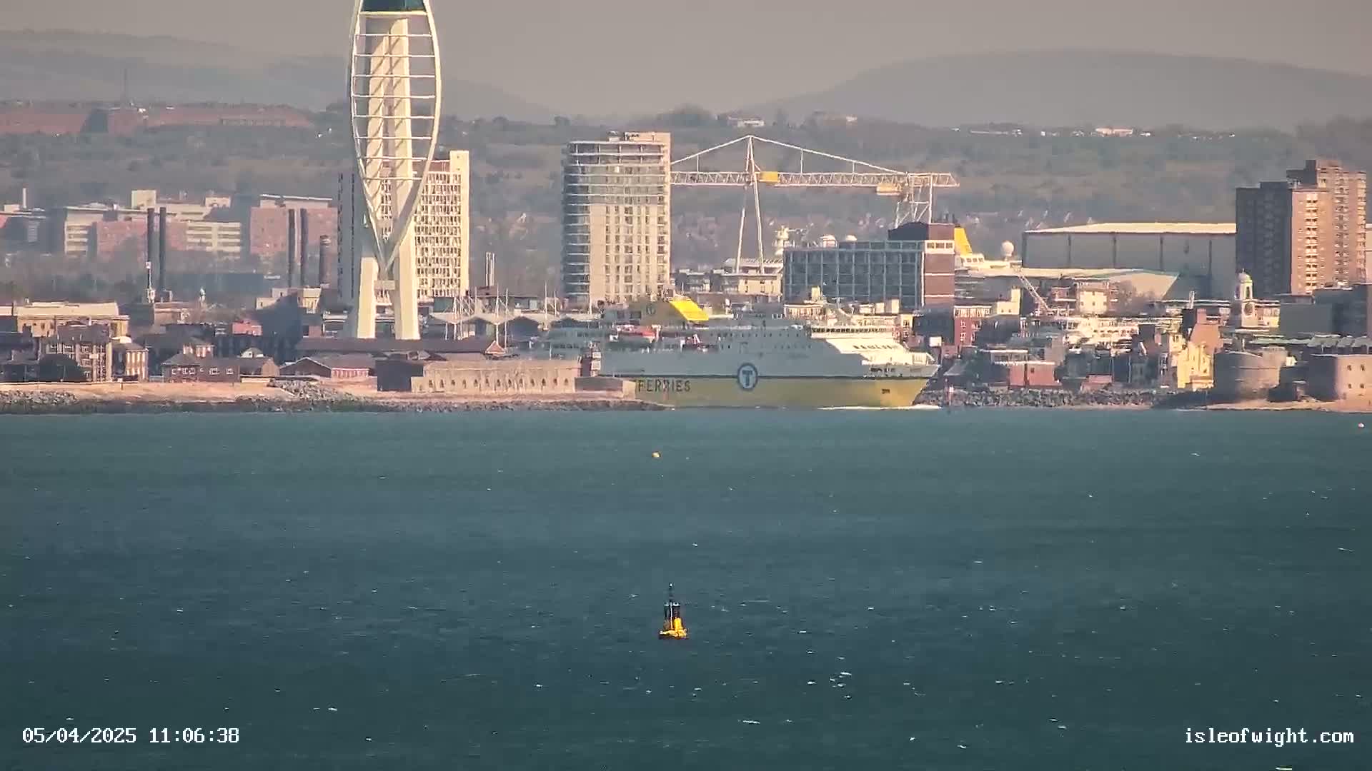 A large yellow ferry is docked near a city skyline on a calm, hazy day.