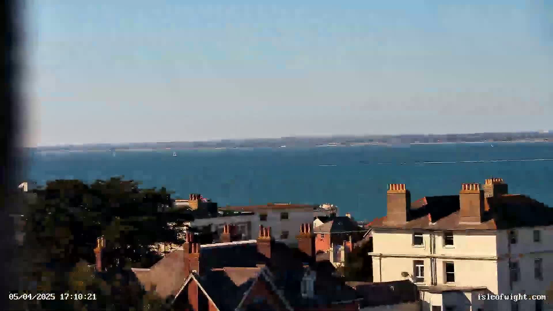 A calm, sunny day overlooks a body of water with several boats visible in the distance, while a collection of houses with brick chimneys are in the foreground.