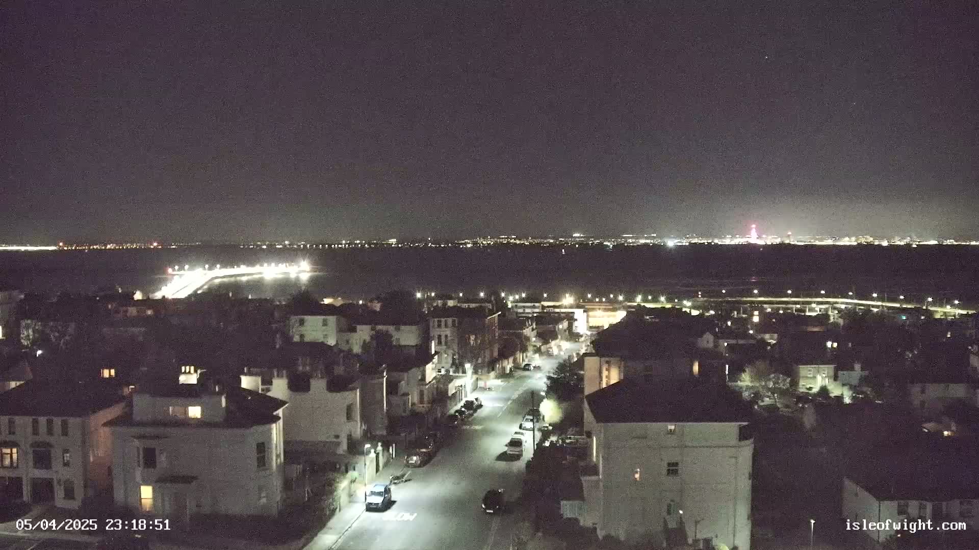 A nighttime view of a residential street with parked cars, overlooking a body of water with distant city lights under a clear, dark sky.