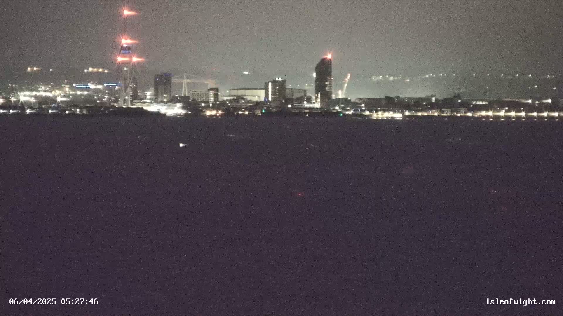 A nighttime view across dark water shows a city skyline with several illuminated buildings, including a tall tower with lights at its top, under a dark, overcast sky.
