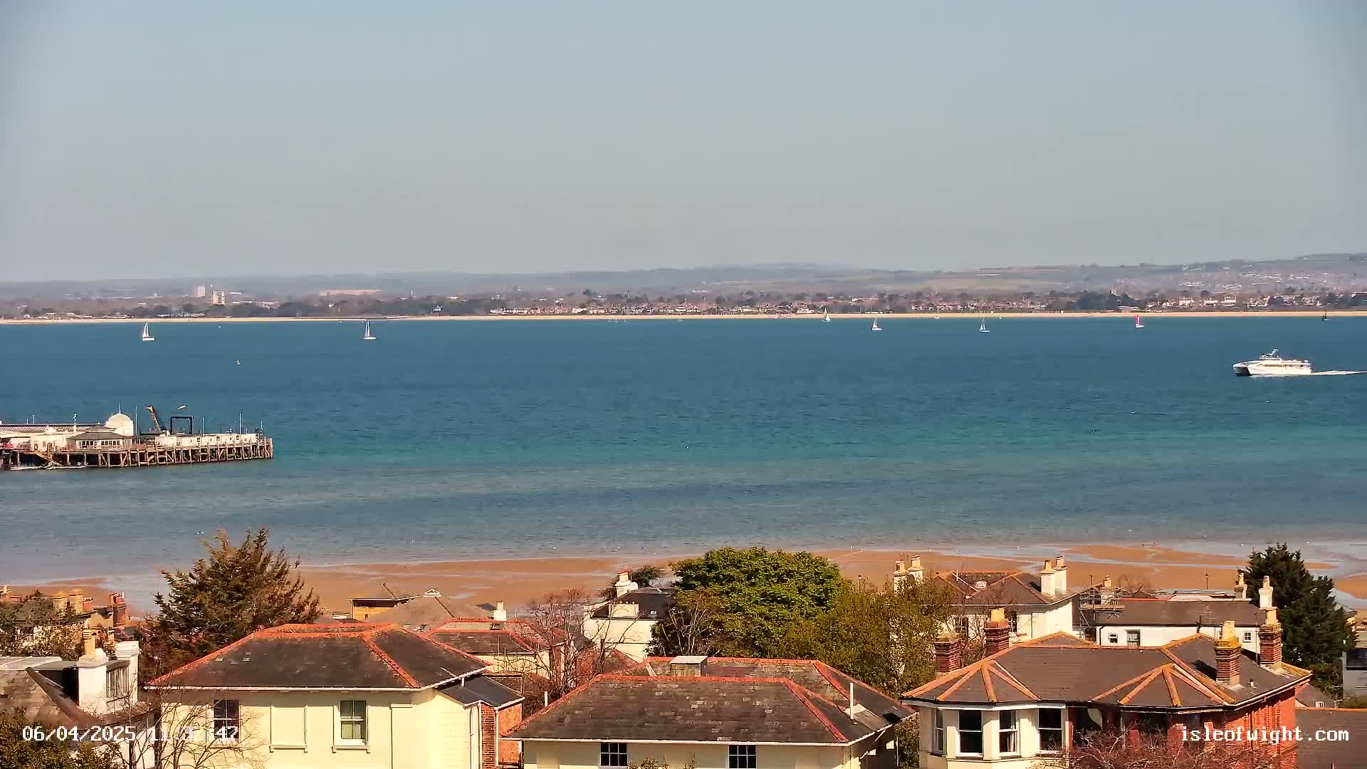 A coastal scene on a sunny day shows a body of water with a pier, several boats, and a distant shoreline, viewed from above a collection of houses with red-tiled roofs.