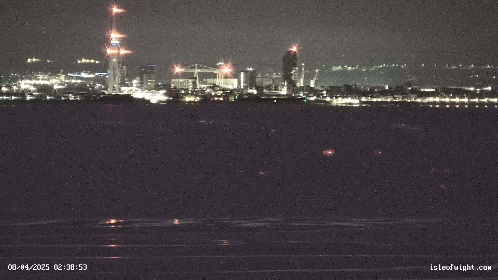 A nighttime view of a city skyline across a dark body of water, with several tall buildings and lights visible.