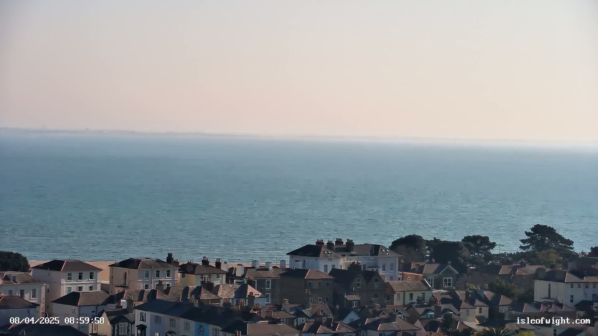 A hazy, sunny day overlooks a calm ocean, with a collection of houses and a small beach visible in the foreground.