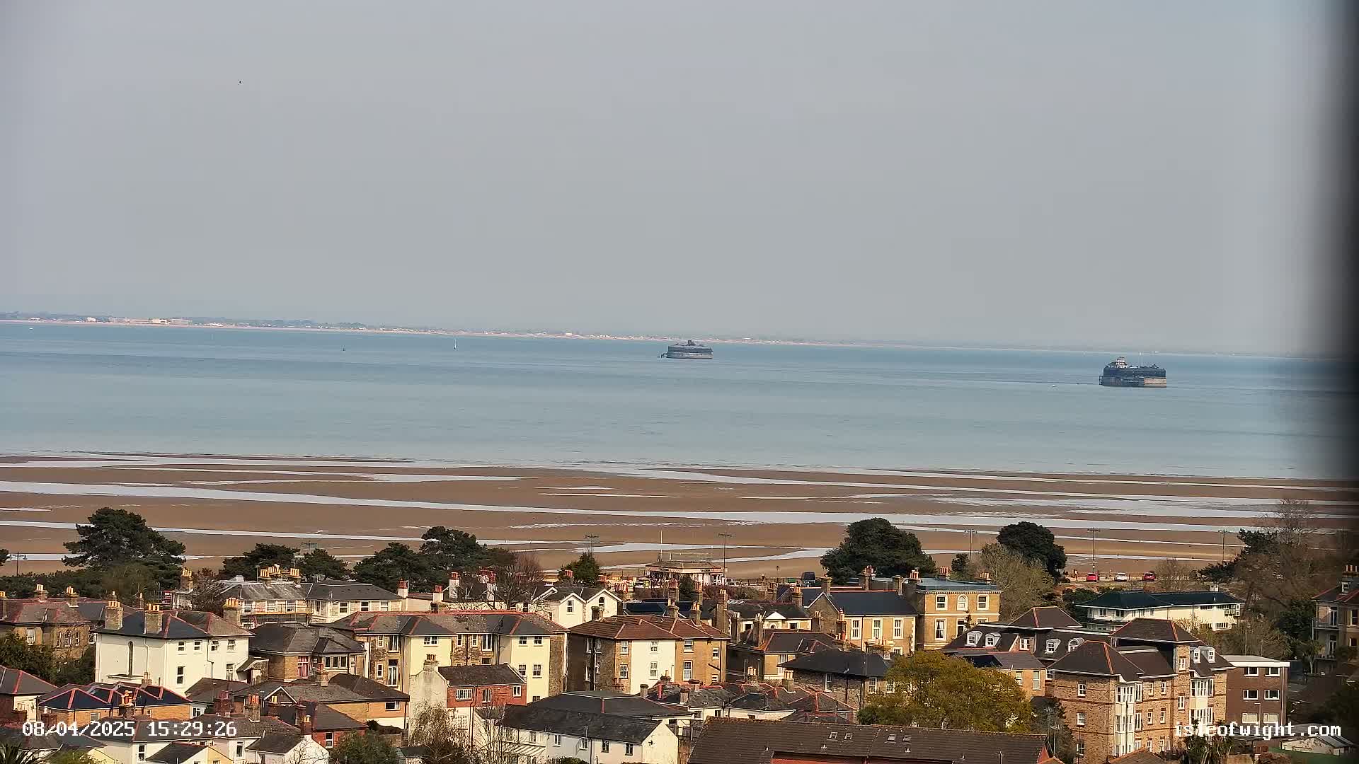 A coastal town overlooks a wide expanse of calm, light-blue water with exposed sandbars at low tide and two distant sea forts visible on the horizon under a hazy sky.