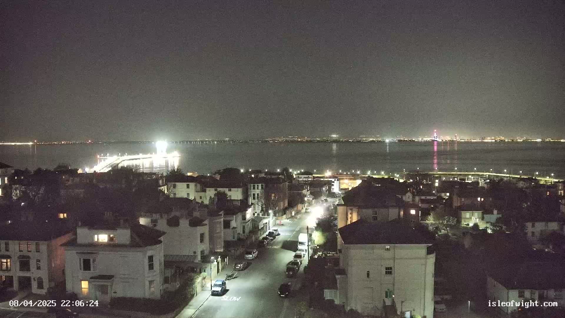A nighttime view of a residential street overlooking a calm body of water with a brightly lit pier and a distant city skyline visible under a clear, dark sky.