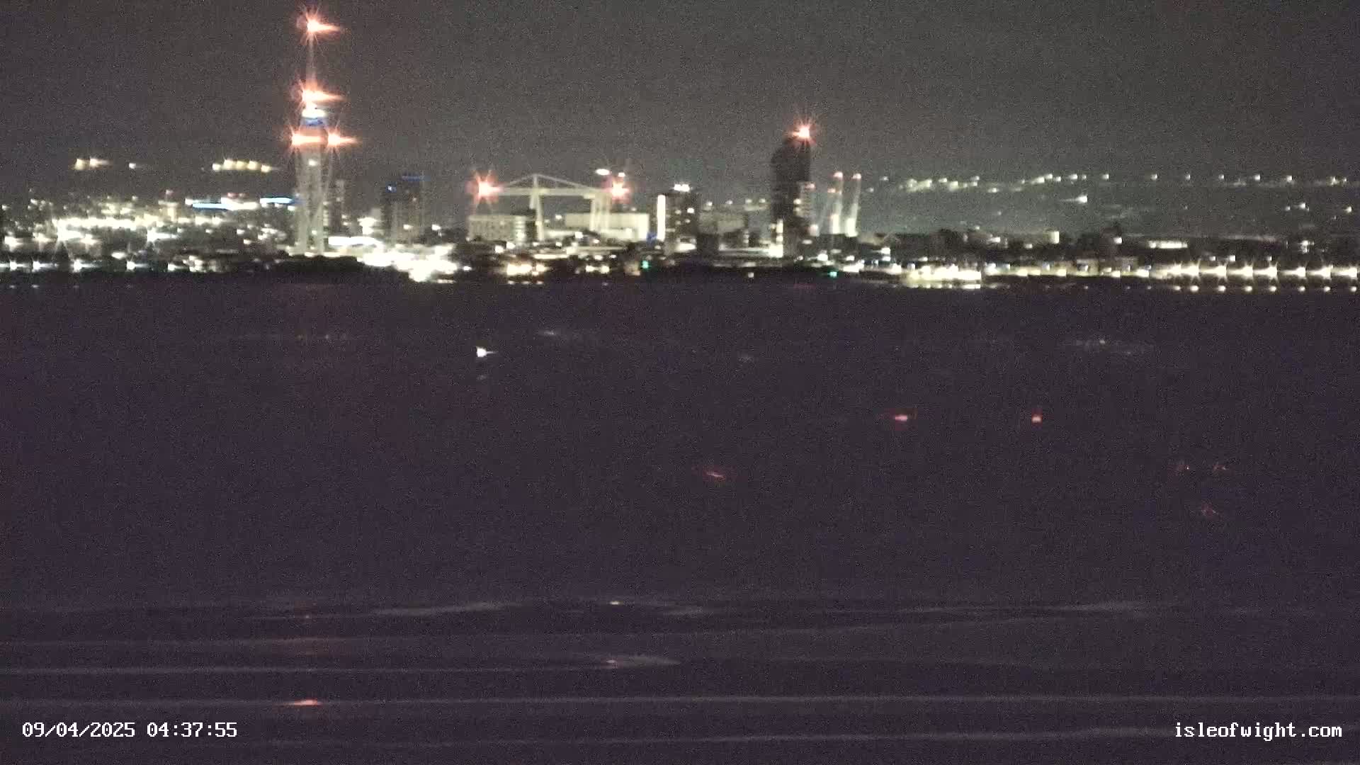 A nighttime view of a city skyline across a dark body of water, with faint ripples visible on the surface.