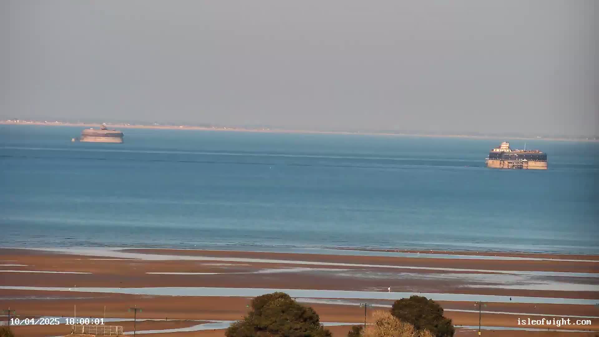 A wide, long shot shows two sea forts in a calm body of water, with a sandy beach receding in the foreground under a hazy sky.
