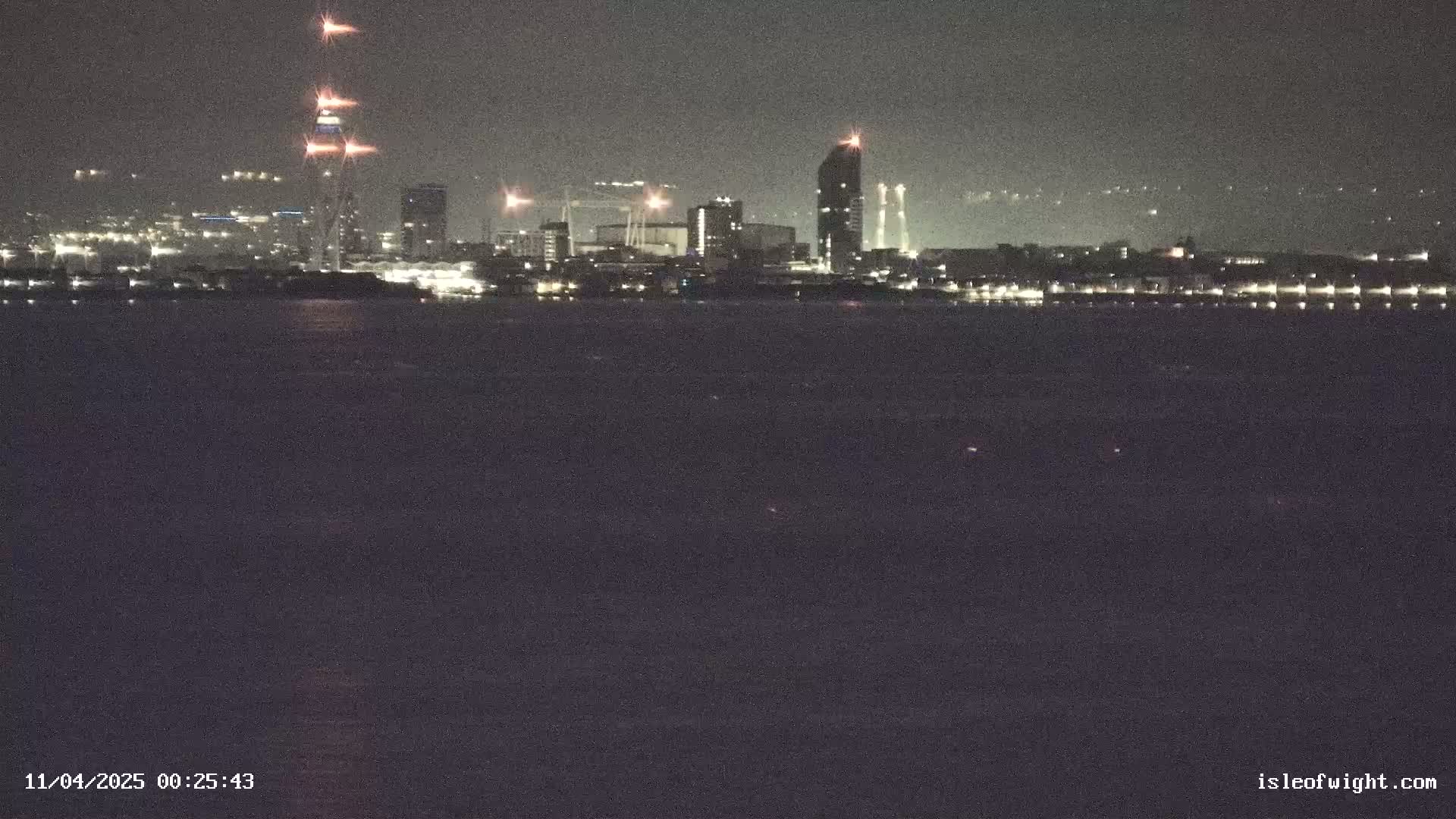 A nighttime view across dark water shows a city skyline with several tall buildings lit up against a dark sky.