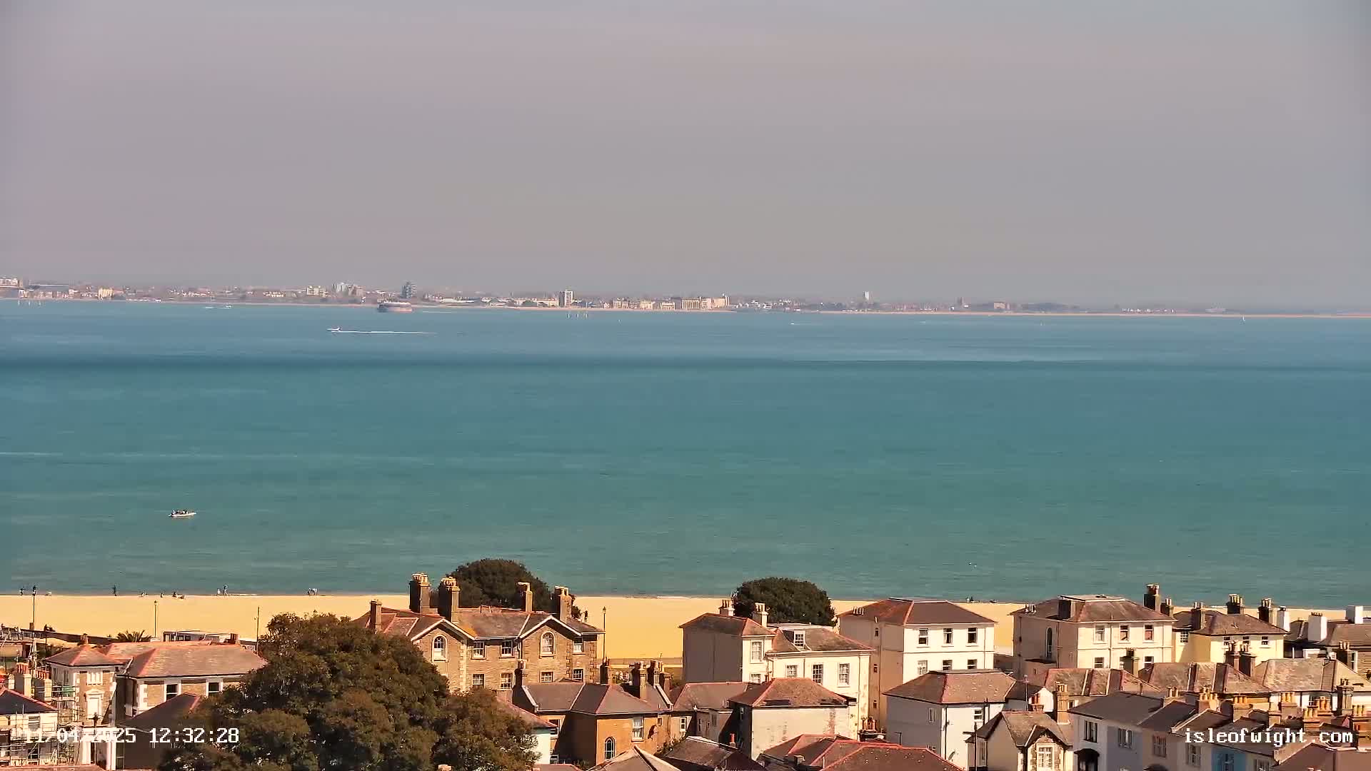 A sandy beach and a row of houses are visible in the foreground, with a calm, teal ocean and a distant city skyline under a hazy sky.