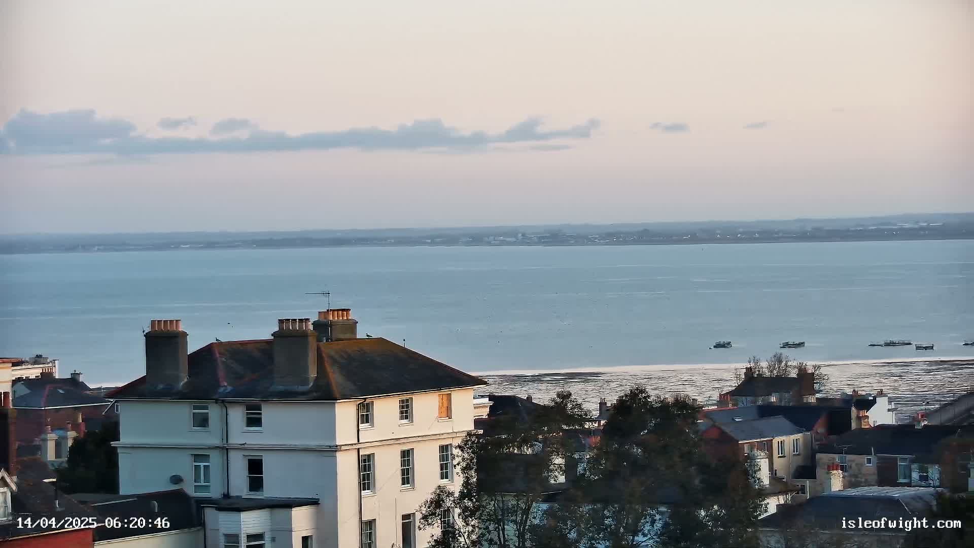 A calm, pale-blue body of water stretches to the horizon under a mostly clear, light-grey sky, with a few small boats visible in the distance, beyond a collection of houses and trees.